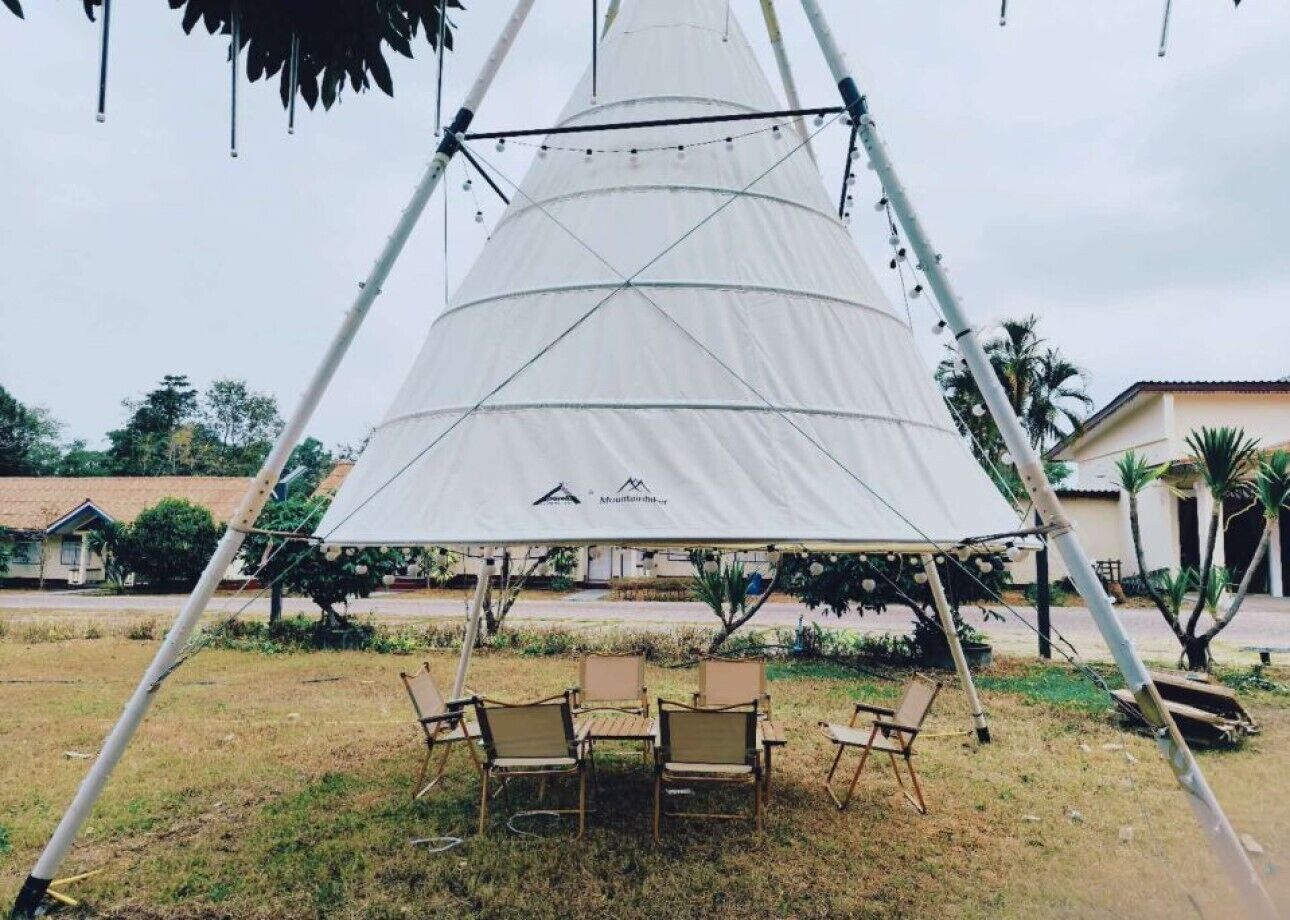 Una carpa blanca grande con un logotipo, rodeada de cuatro sillas y una mesa, montada en un área al aire libre con césped y algunos árboles.