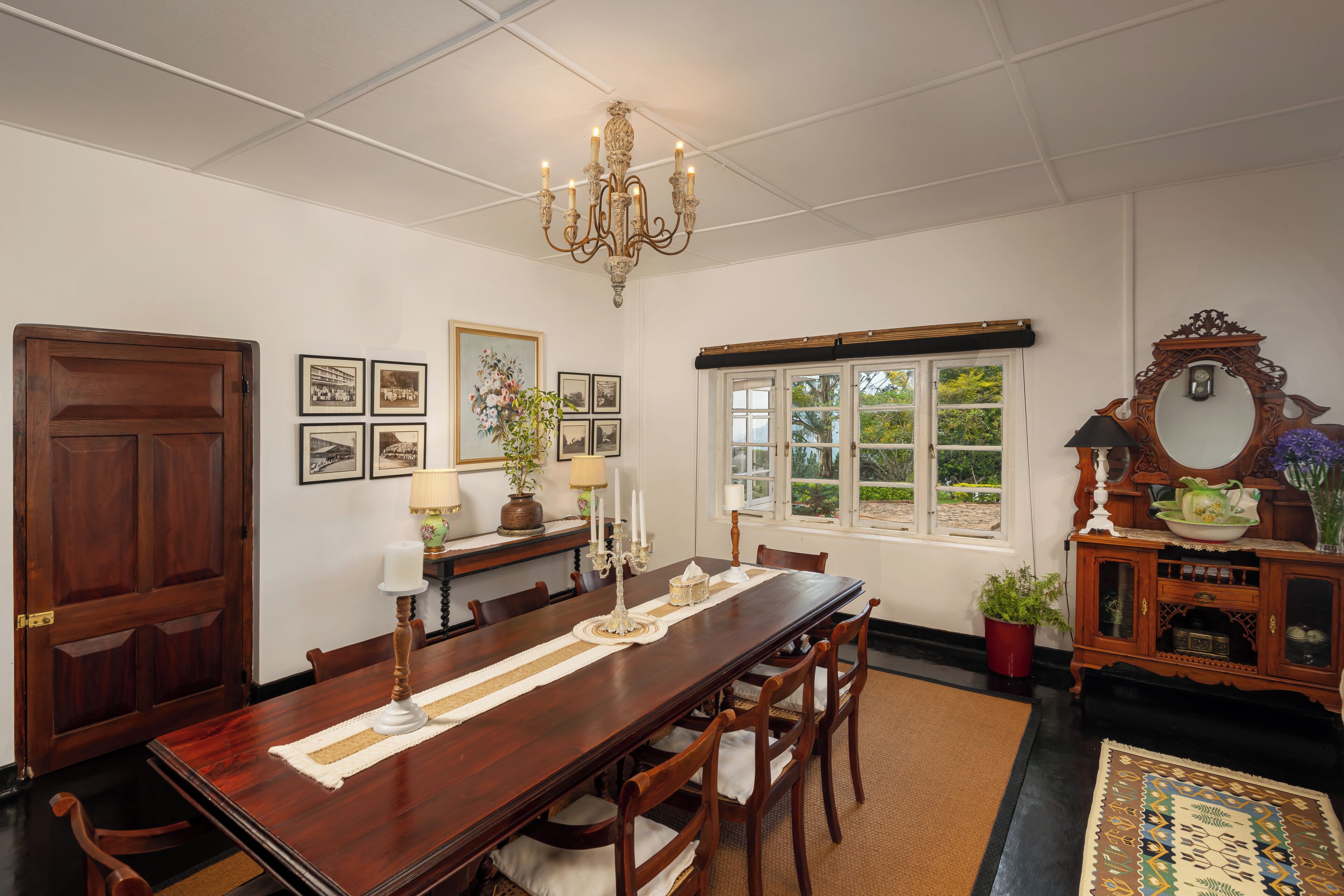 Uma sala de jantar com uma mesa de madeira comprida, cadeiras, um lustre, quadros, uma janela e um espelho.