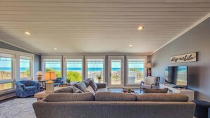 A spacious living room with a large gray sofa, a television, and a view of the ocean through large windows. - The Sandlot - Renovated & Right on the Beach! (Topsail Beach)