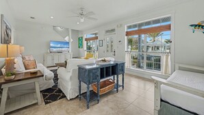 A spacious living area with a white sofa, a blue coffee table, a TV, and a large window with a view of palm trees. - Stairway to Hammock Heaven * Private Pool * Seasonal 2 Beach Sets (Destin)