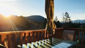 A glass-top table with a wooden railing on a balcony, overlooking a mountainous landscape with the sun setting in the background. - Large Condo Mountain View 5 bed 2.5 bath (Radium Hot Springs)