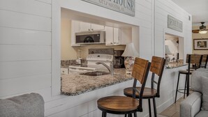 A kitchen with a granite countertop, white cabinets, and a microwave. There are two wooden chairs and a bar stool. A sign above the kitchen reads "Beach House Gulf Shores, AL". - 🌞Low Density | Community Pool | Sea Breeze 203🌞 (Gulf Shores)