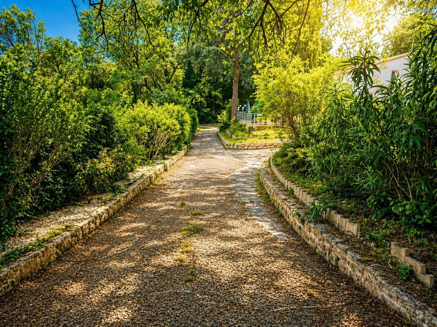 Un sentier bordé de haies soigneusement taillées et d’un mur de pierre, qui mène à un bâtiment à la façade blanche.