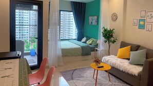 A modern living room with a sofa, a small table, and a chair. There is a bed with a green bedspread and a window with curtains. - SURIHOME- Vinhomes Grand Park Apartments (Ho Chi Minh City)