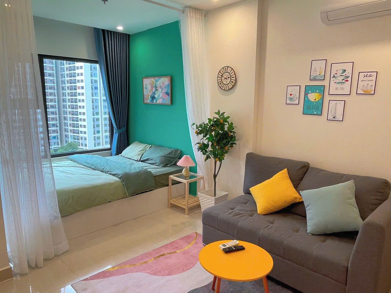 Quarto moderno com cama, sofá cinza, mesa pequena e janela com cortinas.