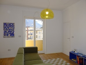 A room with a green sofa, a yellow pendant light, a checkered rug, a bookshelf with books, and a door leading to a balcony.