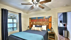 Um quarto com cama, um ventilador de teto, janela com cortinas, cabeceira com obra de arte e uma pequena mesa de cabeceira com abajur.