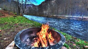 A campfire in a metal bowl with a river and forest in the background.