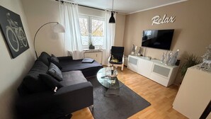 Een moderne woonkamer met een zwarte bank, een glazen salontafel, een televisie en een bordje met de tekst 'Relax'.