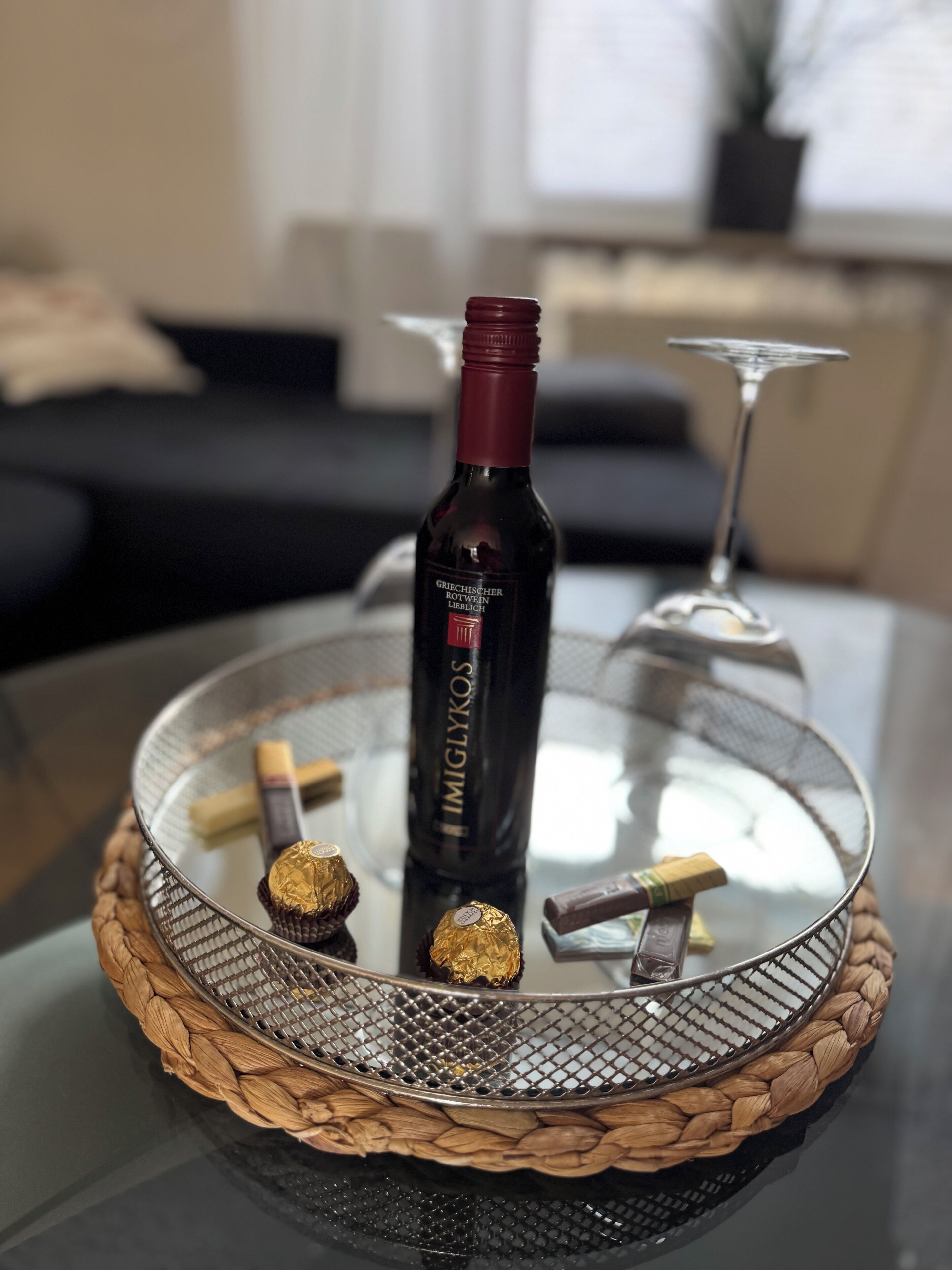 A glass table with a woven base, a bottle of wine, and chocolates.