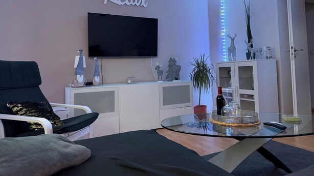 Een woonkamer met een flatscreen televisie, een bank, een glazen salontafel en een kast met decoratieve objecten.