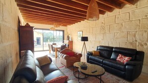 A spacious living area with a black leather sofa, a brown leather couch, and a round coffee table. There is a dining table with chairs, a lamp, and a view through large windows. - CalypSo Luxury Mas Madeleine in Provence (Arles)