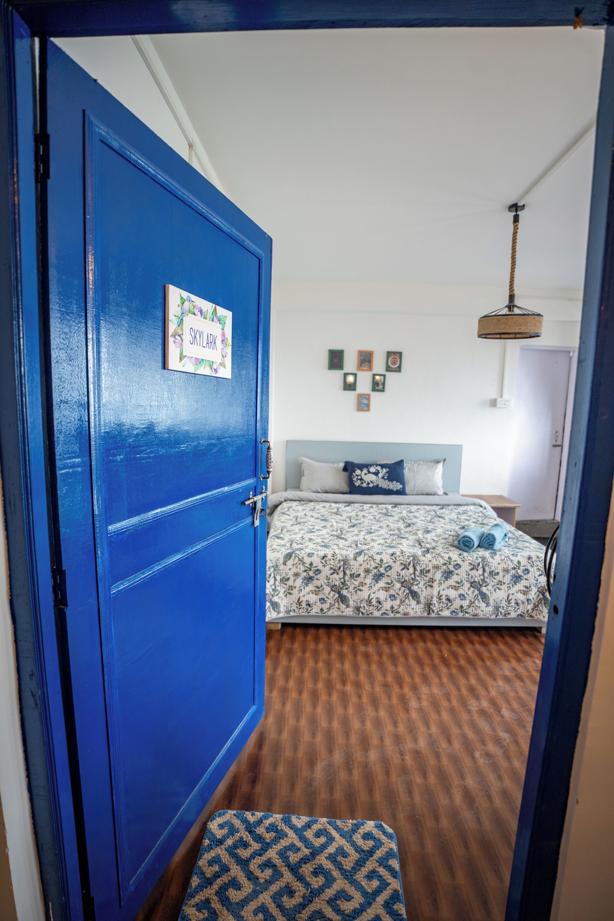 A room with a blue door, a bed with floral bedding, a small rug, and a wall-mounted light fixture.