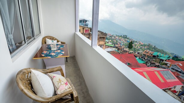 A balcony with a wicker chair, a small table with a floral tablecloth, and a view of a town with colorful buildings and mountains in the distance. - Monratino The Snow roost (Darjeeling)