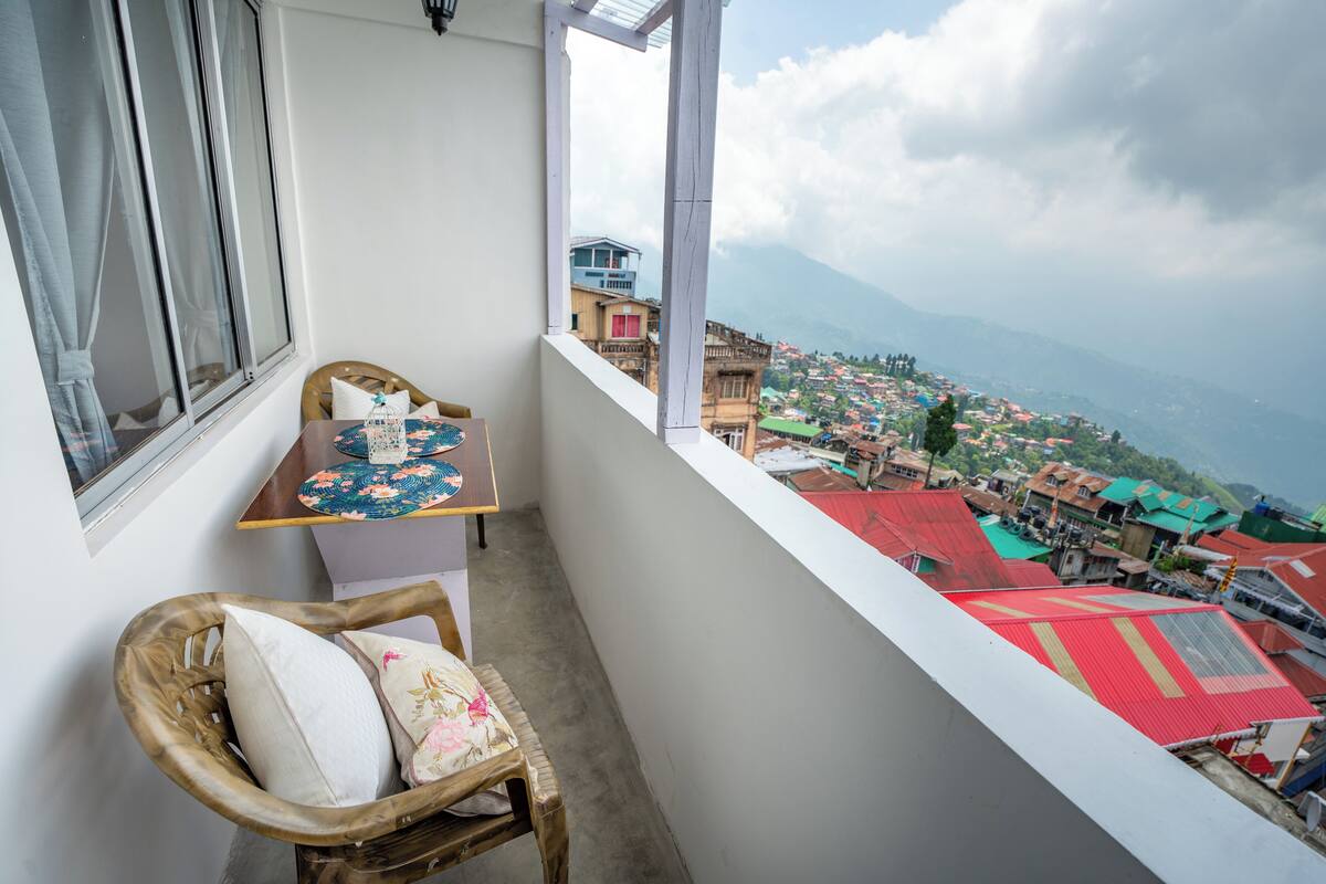 A balcony with a wicker chair, a small table with a floral tablecloth, and a view of a town with colorful buildings and mountains in the distance.