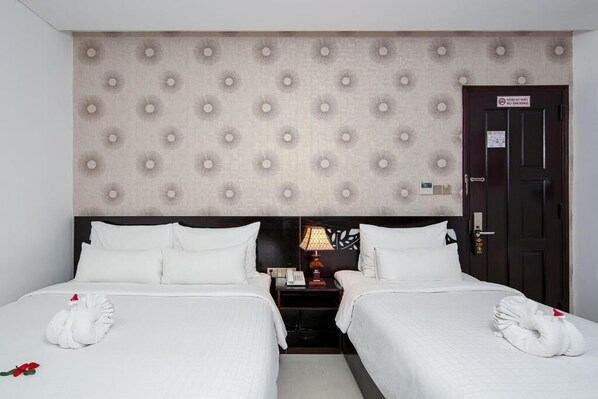 Habitación de hotel con dos camas, cabecera con diseño capitonado, mesita de noche con lámpara y puerta con letrero de 'No Fumar'.