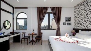 A hotel room with a bed, a desk, a chair, a small table with a bottle and glasses, curtains, a window with a view, and a wall with a patterned design. - Dung Nam Hotel (Da Nang)