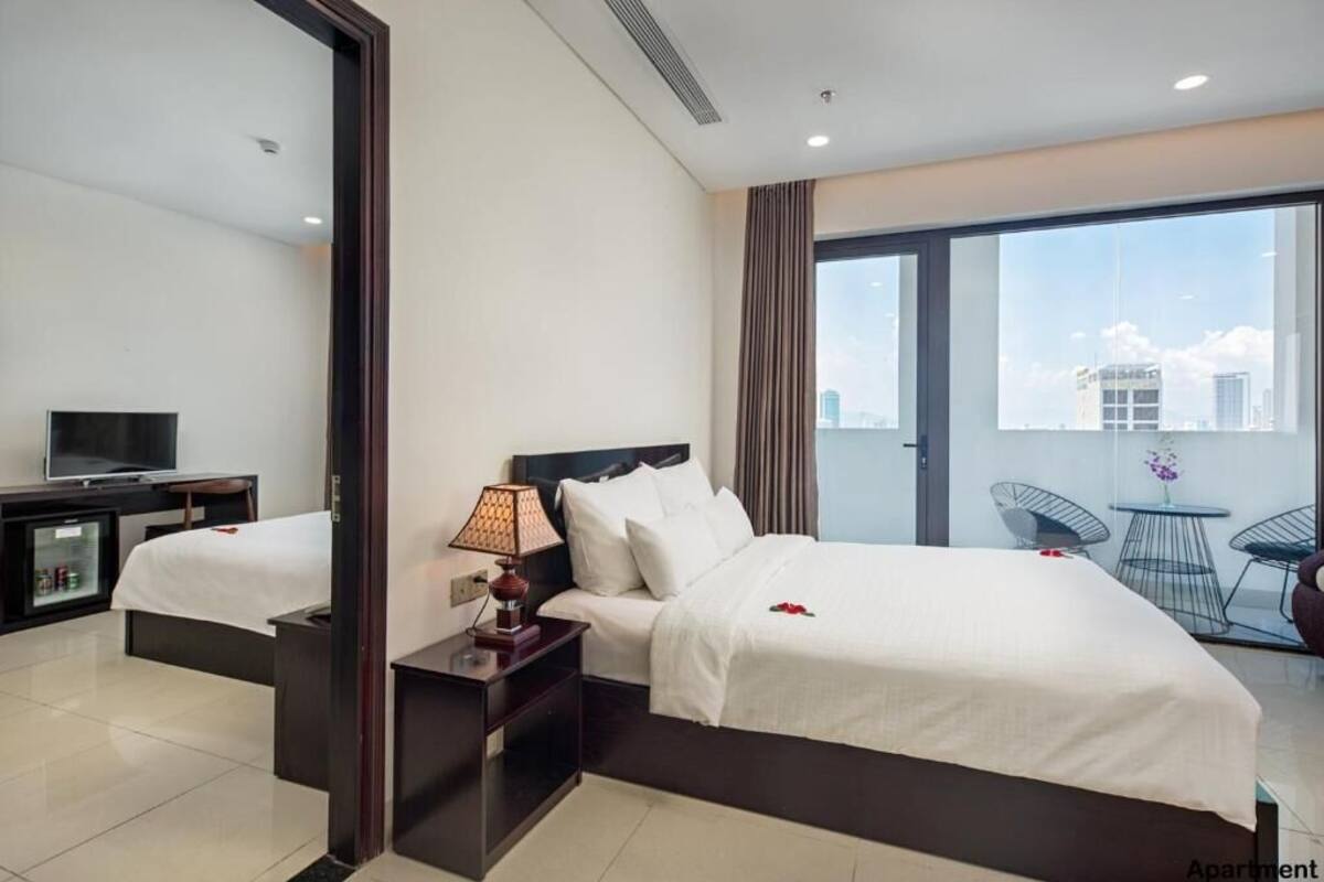 A hotel room with a bed, a TV, a lamp, a small table, and a balcony with chairs.