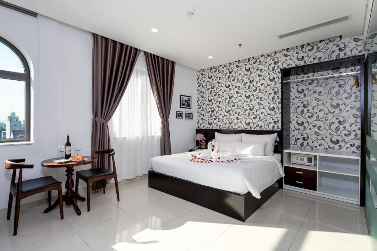 A hotel room with a bed, a small table with a bottle and glasses, a chair, a wardrobe, and a window with curtains.