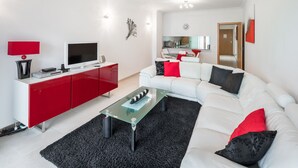 A modern living room with a white sofa, red and black cushions, a glass coffee table, and a red cabinet with a flat-screen TV.