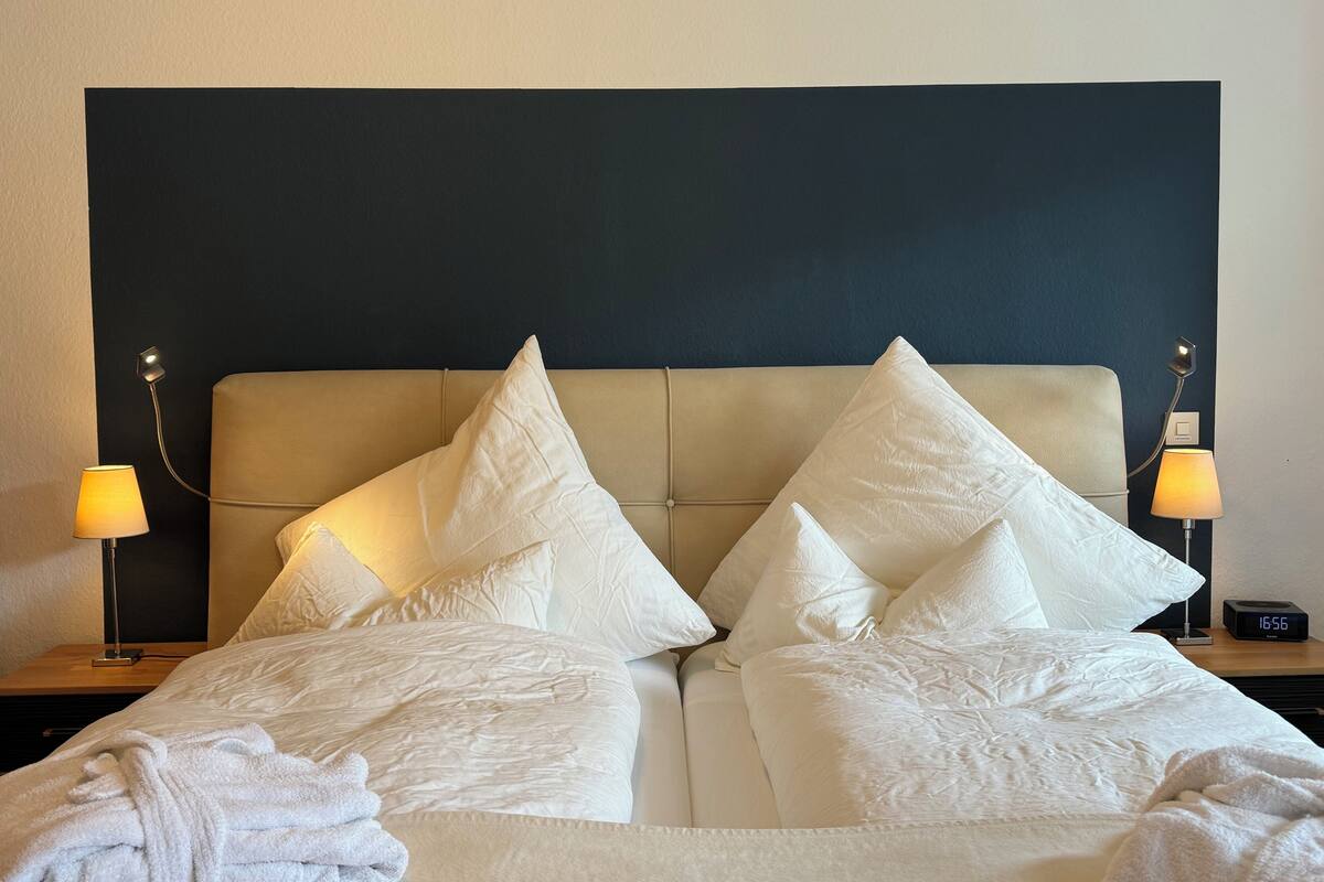 A neatly made bed with white linens and pillows, two bedside lamps, and a digital clock on a bedside table.