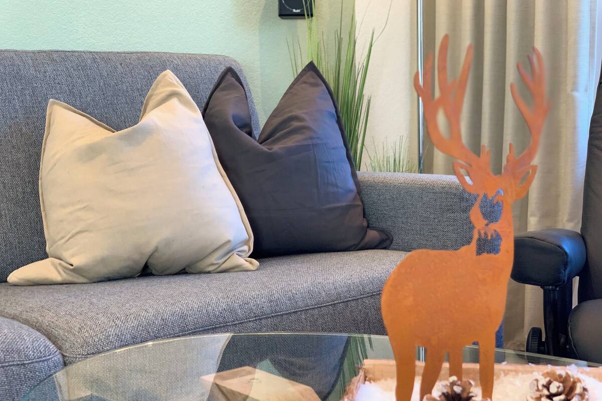 A living room with a grey sofa, beige and dark grey cushions, a glass coffee table with a wooden deer decoration, pine cones, and a plant in the background.