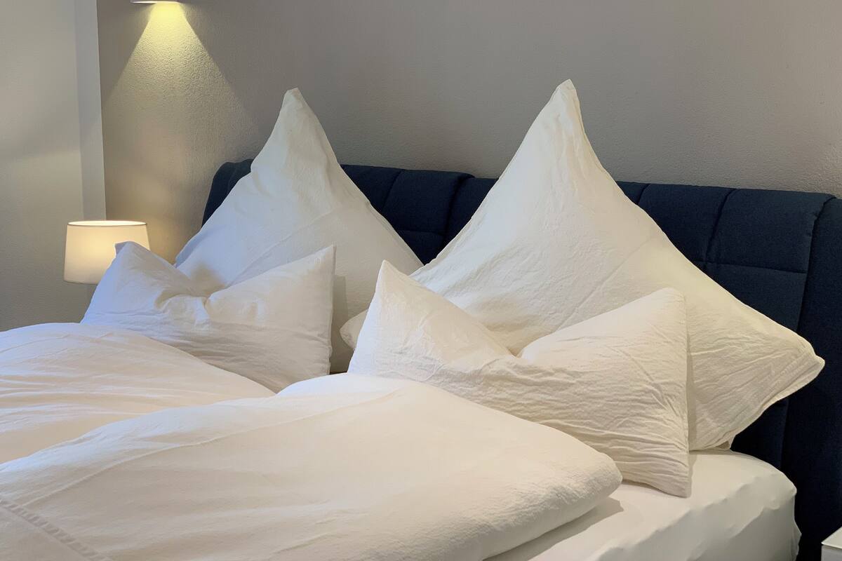 A neatly made bed with white bedding and multiple pillows, a dark blue headboard, and a wall-mounted light fixture.