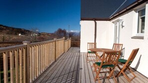 Una terraza de madera con una mesa y sillas, un edificio blanco con techo oscuro, y un cielo azul despejado.