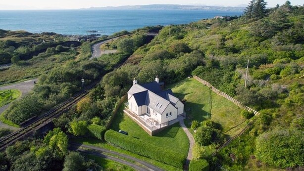 Una casa grande de un solo piso con techo pronunciado, rodeada de abundante vegetación y vistas al mar.