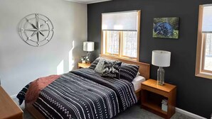 A bedroom with a bed, two bedside tables, a wall clock, a window with blinds, and a framed picture.