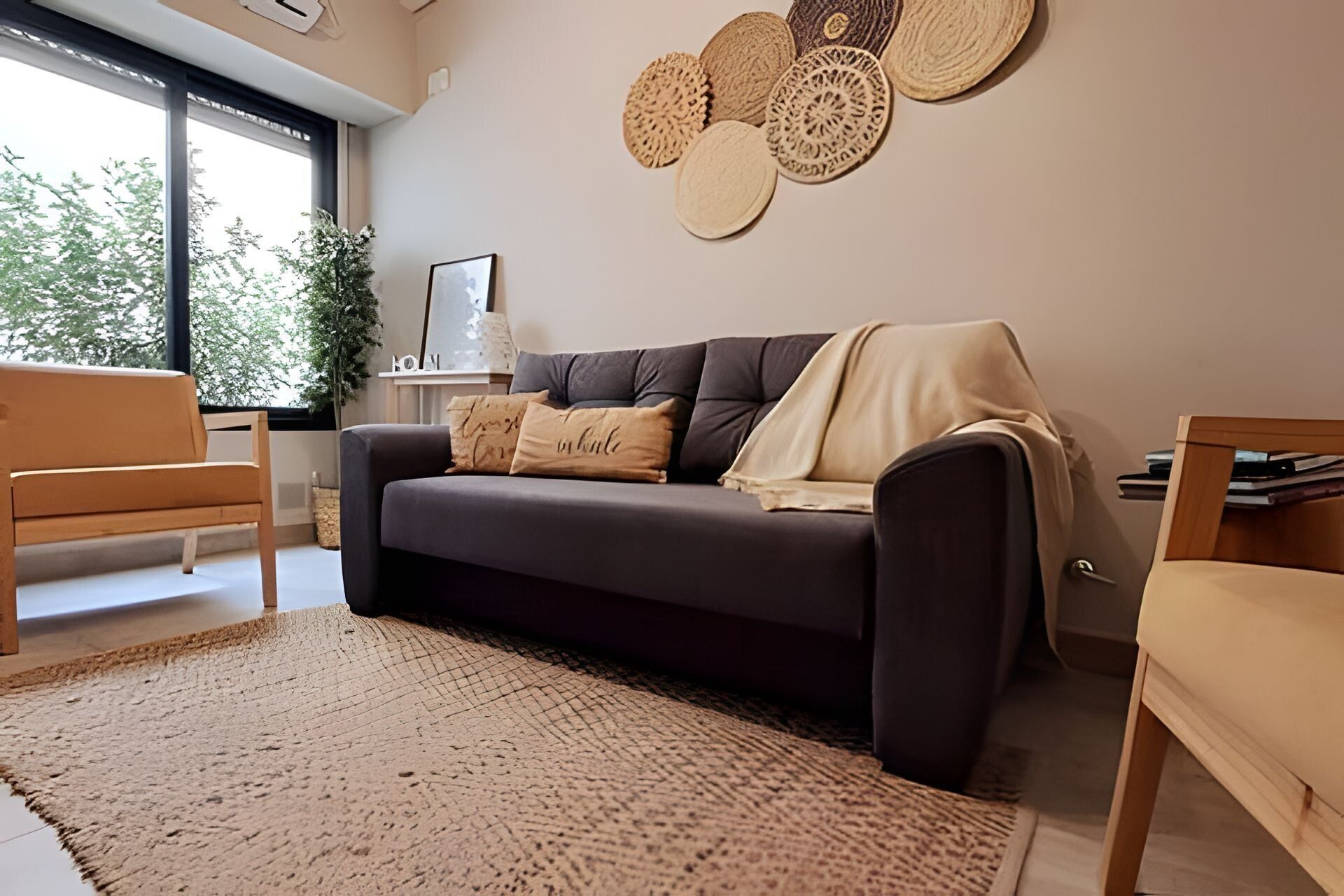 Una sala moderna con un sofá gris oscuro, dos sillas de madera, un tapete tejido grande y una pared decorada con piezas de arte tejidas circulares.
