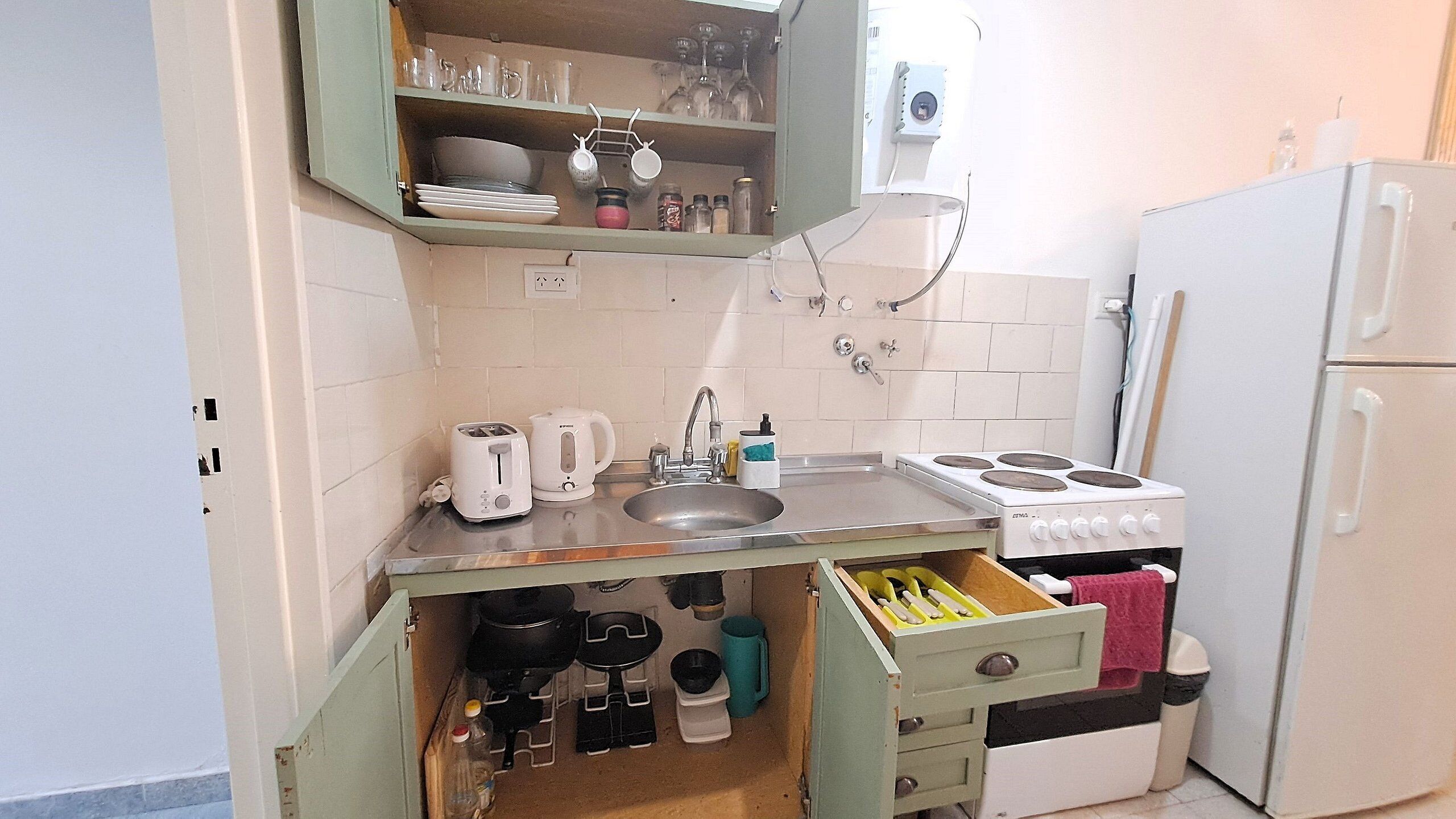 Una cocina compacta con un lavamanos, una estufa y un refrigerador. Hay armarios sobre el lavamanos y una encimera con un tostador y un hervidor.
