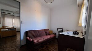 A small, sparsely furnished room with a wooden floor, a striped sofa, a wooden bed, a small table with papers, and a chair. - Hornera Mai - The perfect place (Rosario)