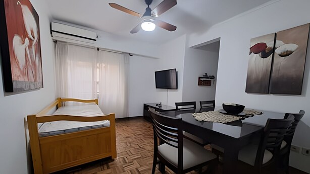 A small, well-lit room with a bed, a dining table with chairs, a ceiling fan, a flat-screen TV, and artwork on the walls. - Hornera Mai - The perfect place (Rosario)