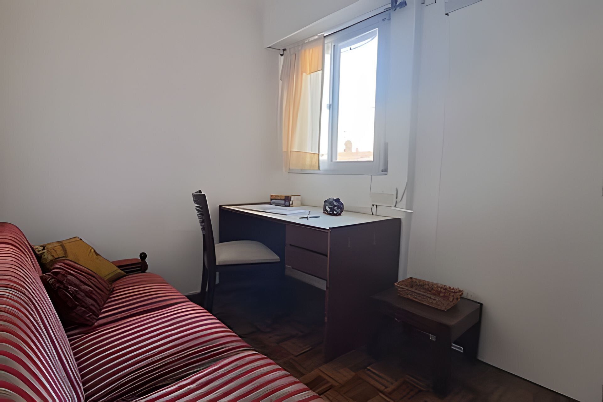A small, sparsely furnished room with a single bed, a desk, and a window.