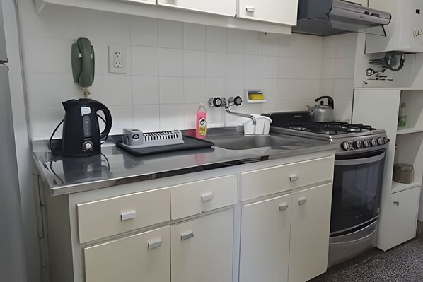 A kitchen with a stove, kettle, and a coffee maker. There is a white cabinet with drawers and a countertop.
