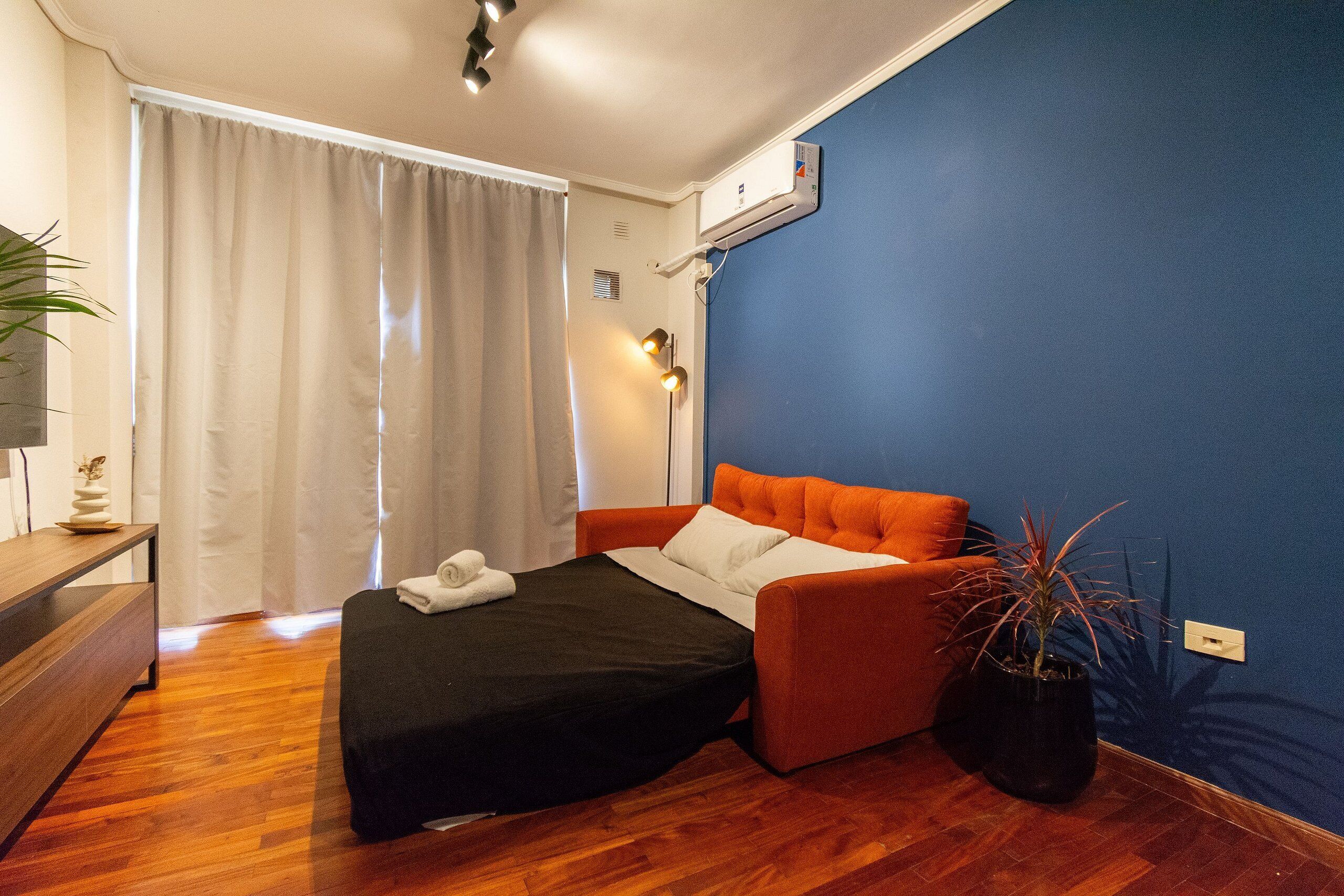 Quarto com cama, mesa de cabeceira, planta em vaso e janela com cortinas.