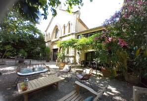 Ein Außenbereich mit einer Terrasse, Korbmöbeln, einem Tisch mit Getränken und einem Gebäude mit Rundbogenfenstern im Hintergrund.