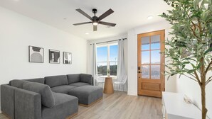 Ein modernes Wohnzimmer mit einem grauen Ecksofa, einem Deckenventilator, einer Holztür und einem großen Fenster mit Vorhängen.