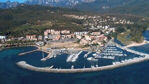 An aerial view of a coastal town with a marina, residential buildings, and mountainous terrain in the background.