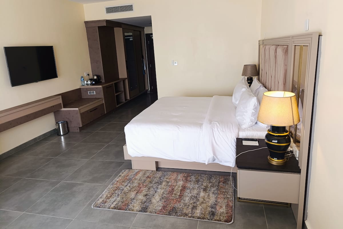 Une chambre d’hôtel comprenant un lit, une table de chevet, une lampe, une télévision et un petit coin salon avec un banc et des rangements.