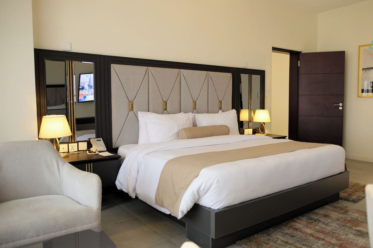 Une chambre d’hôtel avec un grand lit, deux tables de chevet avec des lampes, une chaise, une porte sombre et un tableau encadré au mur.