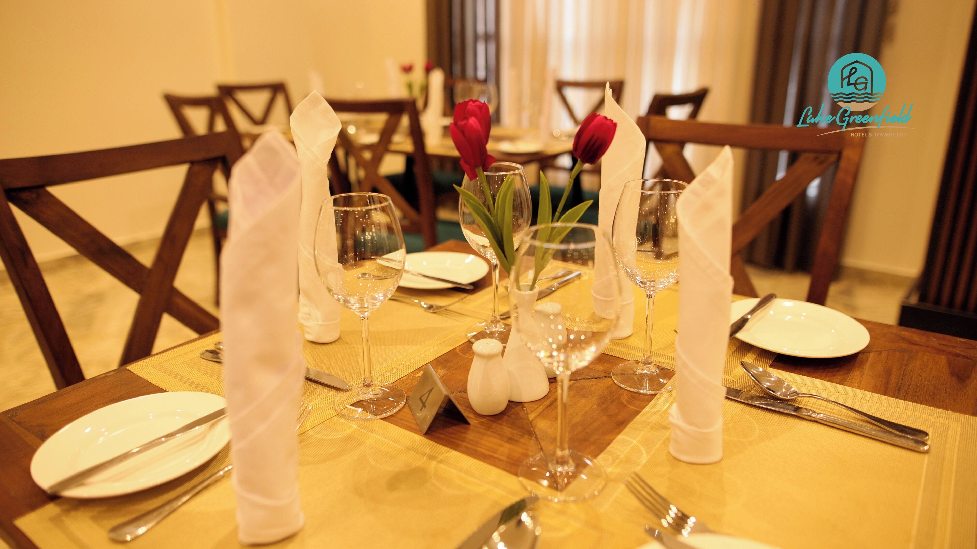 Une table à manger dressée pour quatre personnes, avec des serviettes blanches, des verres à vin et un vase rempli de fleurs rouges.
