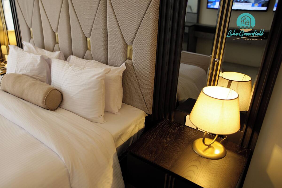 Un lit bien fait, avec du linge de lit blanc et une tête de lit beige, agrémenté de deux lampes de chevet et d’un miroir qui reflète la pièce.