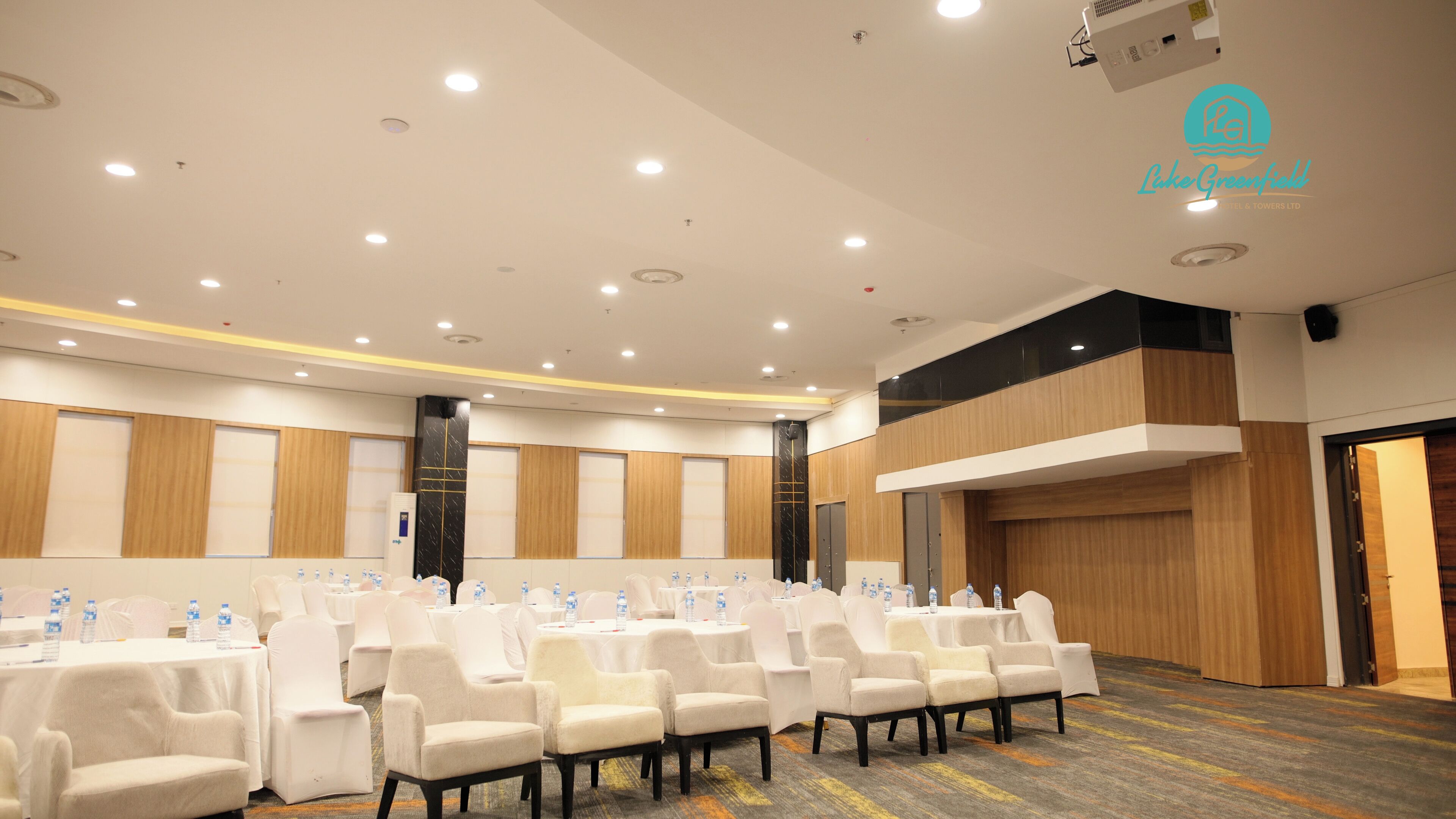 A conference room with rows of white chairs, tables with water bottles, and a modern ceiling with recessed lighting.