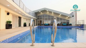 Una piscina interior con agua azul cristalina, rodeada por un solárium con baldosas. Se observan dos escaleras metálicas en primer plano. La zona de la piscina está al lado de un edificio con amplios ventanales y una zona de estar cubierta con sillas azules.