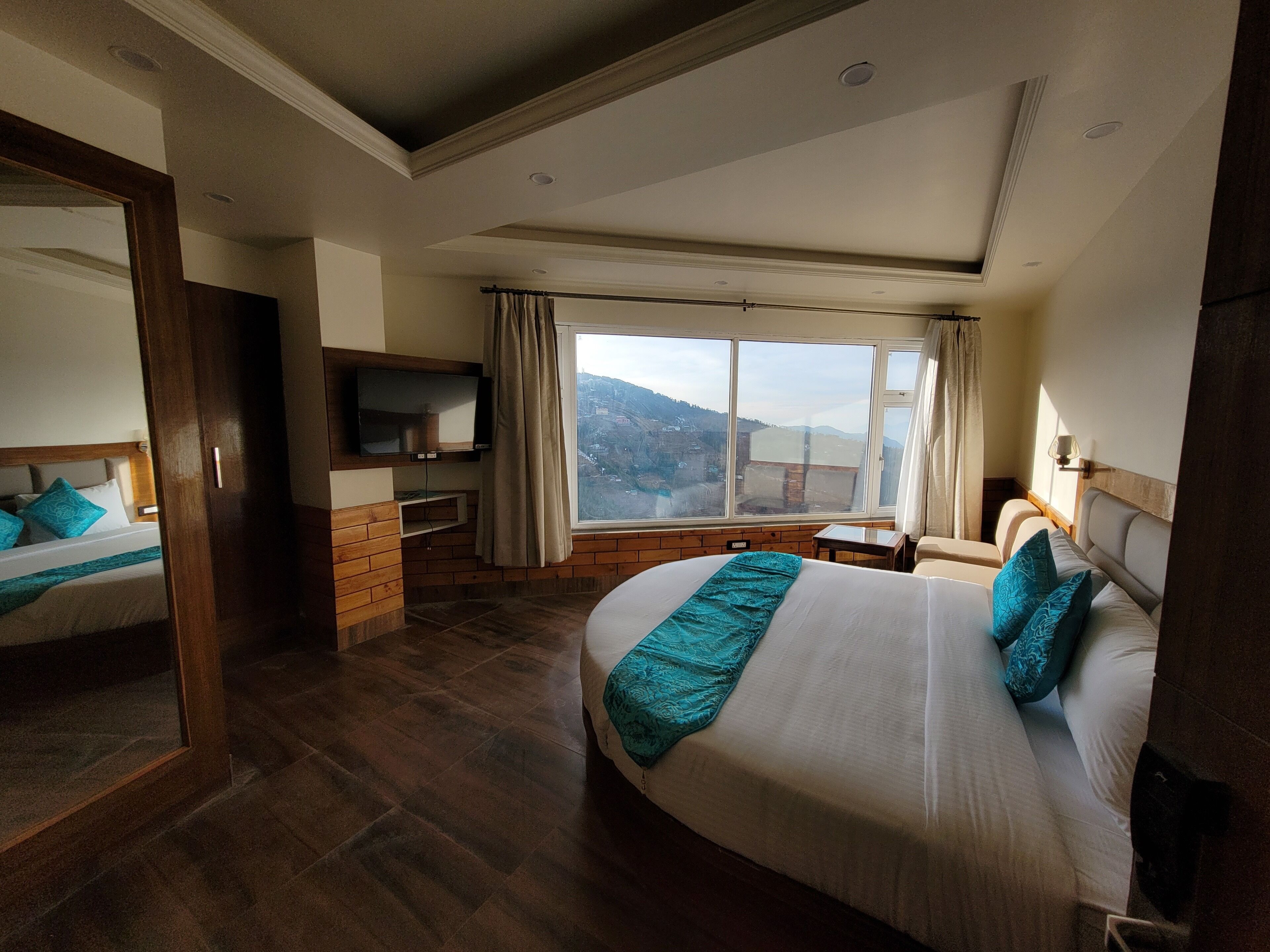 A hotel room with a large bed, a TV mounted on the wall, a small table, and a view of the mountains.
