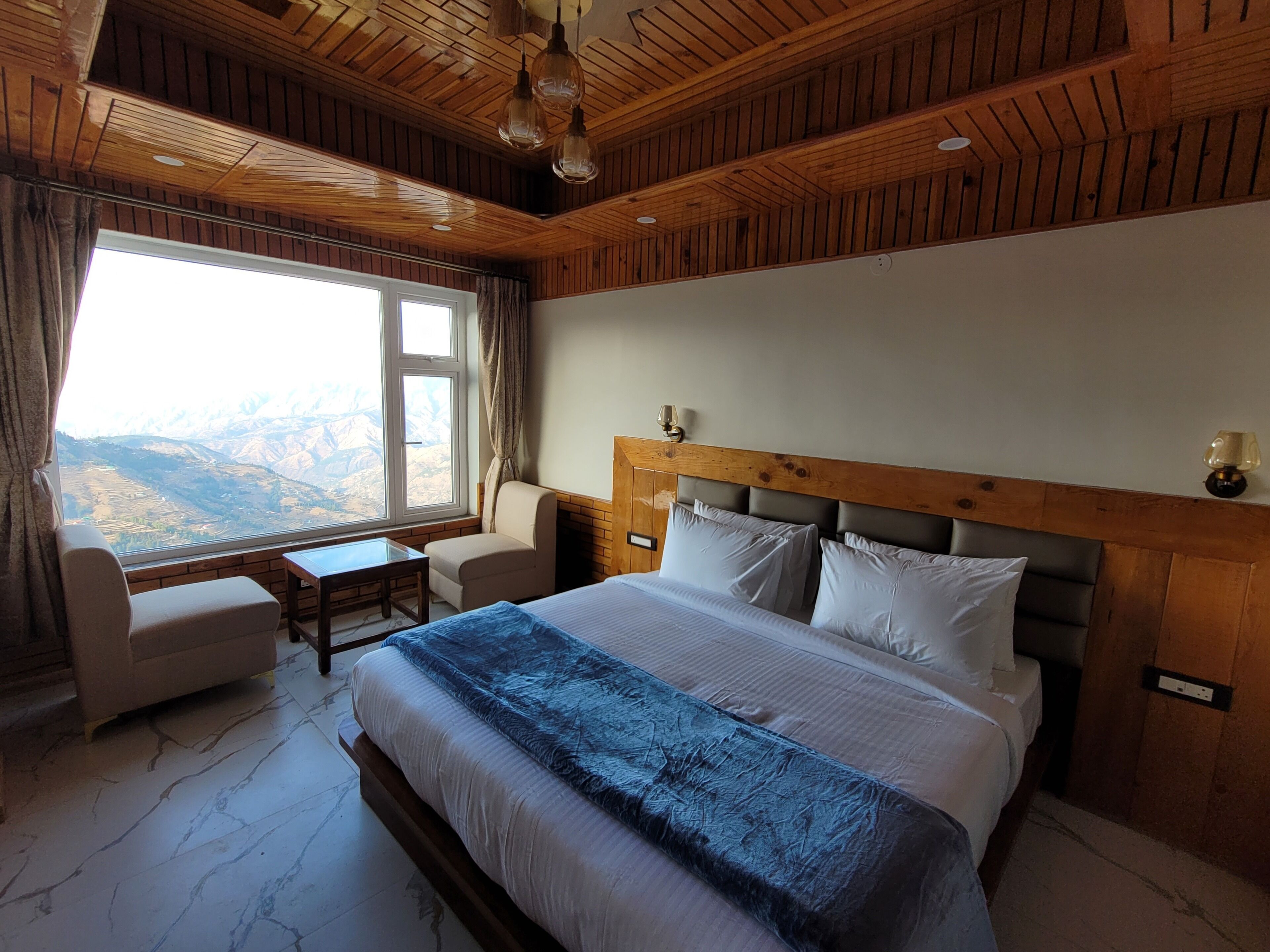 Une chambre avec un grand lit, une tête de lit en bois, une table de chevet, une chaise, une table et une fenêtre donnant sur la montagne.