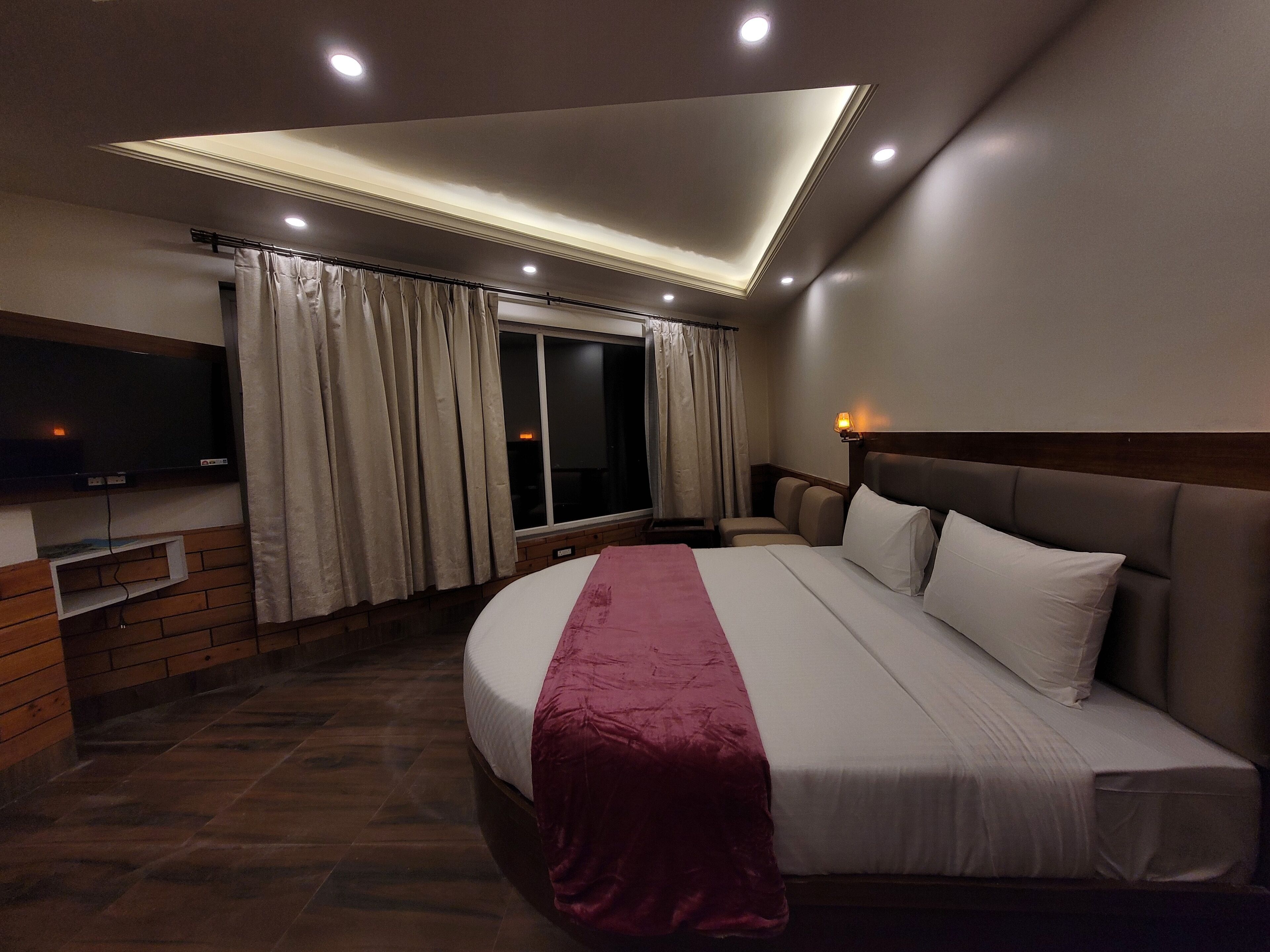 A hotel room with a large bed, a television mounted on the wall, a window with curtains, and a ceiling light fixture.