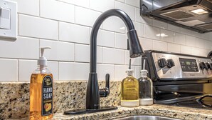 Una cocina con un lavamanos de acero inoxidable, un grifo negro, una botella de jabón para manos y una botella de aceite sobre la encimera de granito.